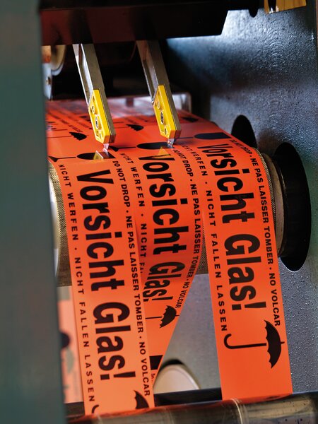 Das Bild zeigt, wie individuell bedruckte Bänder der IBZ zugeschnitten werden. Die Bänder sind orange und in der Maschine eingespannt. Auf den Bändern steht Vorsicht Glas!